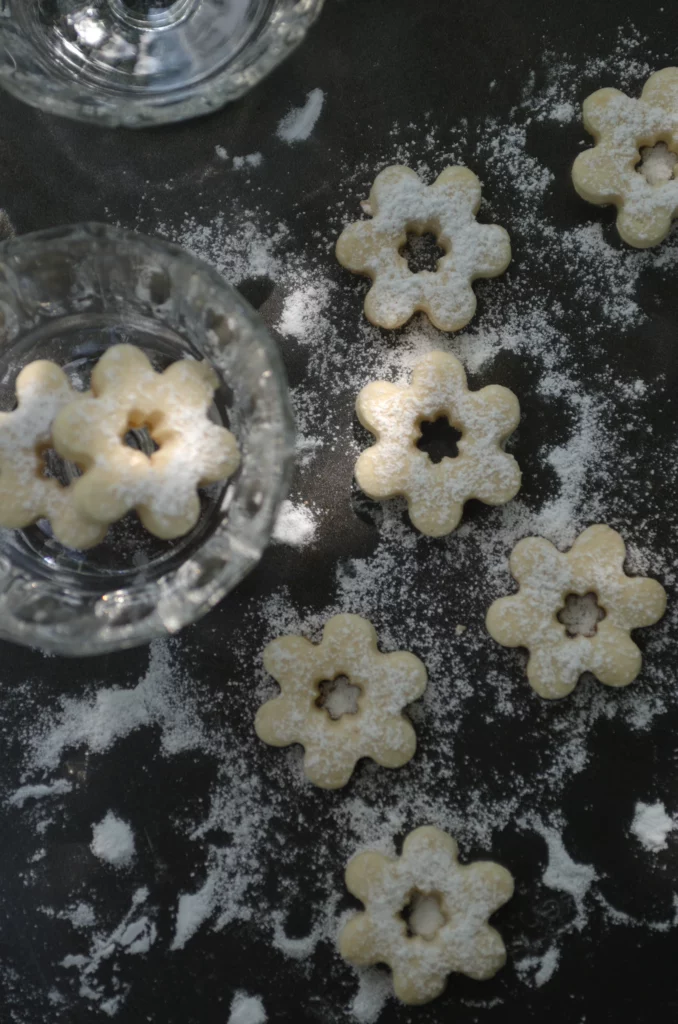 Several Italian vegan Canestrelli biscotti on the black, metal surface, laying in the shadow of a tree above. A glass vase contains two of Canestrelli biscuits. Biscuits are covered by white icing sugar.